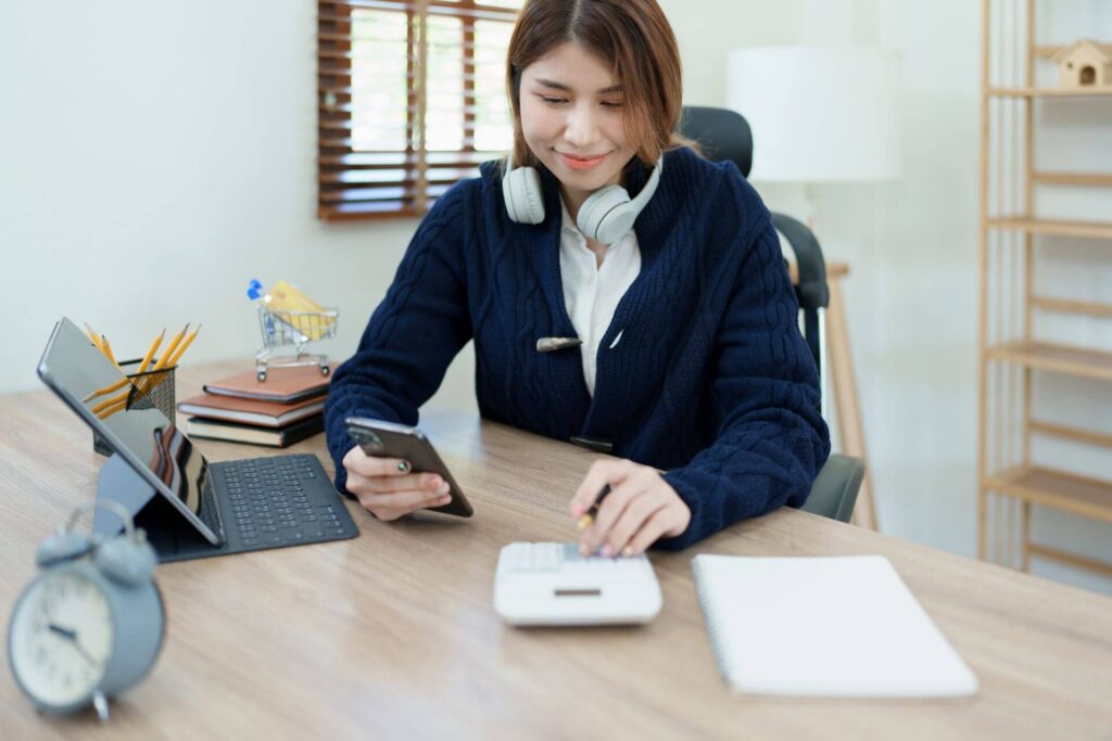 Person wearing headphones paying car loans responsibly at desk via smartphone and calculator, with piggybank with clock in foreground